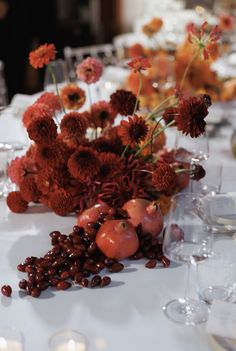 an arrangement of flowers and fruit on a table with wine glasses in the foreground