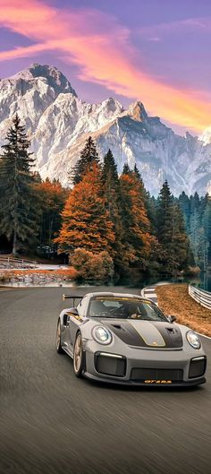 a grey sports car driving down a road with mountains in the background and trees on both sides