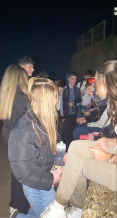 a group of young people sitting around each other on the beach at night, talking and drinking