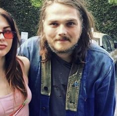 a man with long hair standing next to a woman in a pink shirt and jean jacket