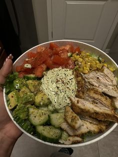 a person holding a bowl filled with chicken, cucumber, tomatoes and avocado