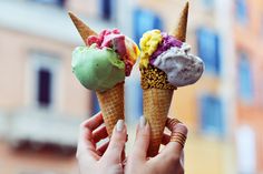 two ice cream cones with colorful toppings being held up in front of a building