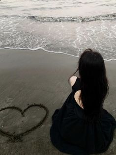 a woman sitting on the beach with her back to the camera and drawing a heart in the sand