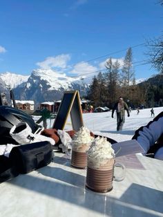 two cups of hot chocolate and whipped cream sit on a table in front of snow covered mountains