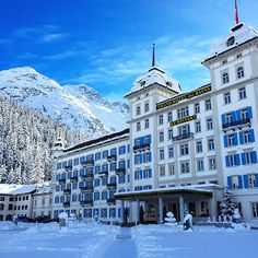 a large white building with blue shutters in front of snow covered mountains and trees