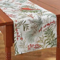 a wooden table topped with a white and red table runner covered in holly leaves, berries and pine cones