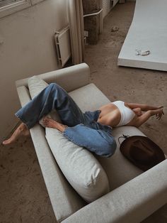 a woman laying on top of a white couch next to a brown hat and window