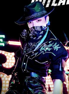 a man wearing a black hat and leather pants with tattoos on his face is standing in front of a neon sign