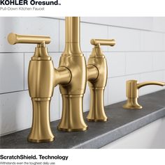 three faucets are shown in gold on a counter top with white tiles behind them