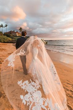 a bride and groom walking on the beach with their wedding veil blowing in the wind