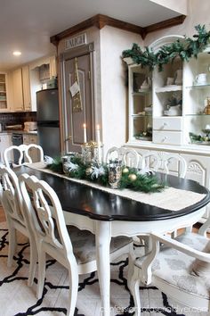 a dining room table decorated for christmas with greenery and candles on it, surrounded by white chairs