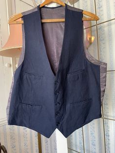 This vest is made of a cotton blend in classic dark navy blue, with a silky dark grey blue back. It has the original buttons in the front, four front pockets  and is fully lined. There are no labels. The measurements, taken with the vest lying flat, are: shoulder to shoulder, 12 inches; armpit to armpit, 21 1/2 inches; length, 24 1/2 inches in front and 21 inches in back; bottom edge, 20 inches. In  very good condition. Formal Sleeveless Cotton Outerwear, Blue Cotton Vest With Buttons, Navy Sleeveless Outerwear With Pockets, Navy Sleeveless Vest With Pockets, Fitted Navy Sleeveless Vest, Classic Fitted Cotton Denim Vest, Classic Fitted Navy Vest, Fitted Classic Cotton Denim Vest, Navy Fitted Vest For Workwear