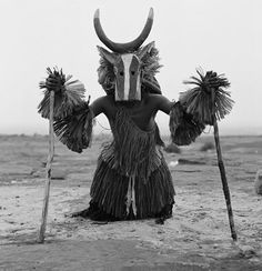 a person with long hair and horns standing in the desert holding two sticks, wearing an animal mask