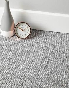 a clock sitting on the floor next to a gray vase and white wall with grey carpet