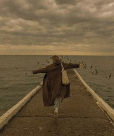 a woman is walking down a pier towards the ocean with birds flying around and over her