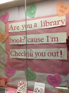 a library display with pink and white paper hearts on the wall, reading are you a library book? because i'm checkin'm out