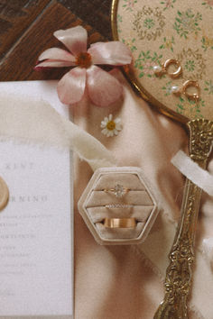 wedding rings and ring box on table next to mirror with flower in it, along with other items