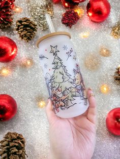 a hand holding a tumbler with a christmas scene on it next to ornaments and pine cones