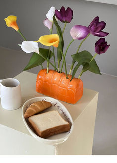 an arrangement of flowers in a vase with bread and coffee on a table next to it