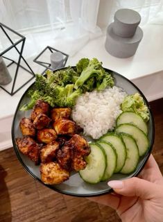 a person holding a plate with chicken, broccoli and rice on it in front of a window