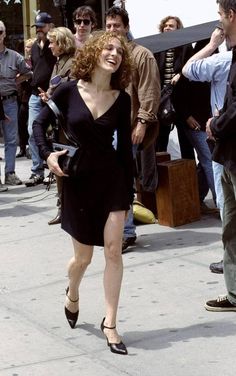 a woman in black dress walking down the street with people behind her and onlookers