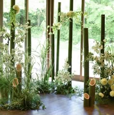 an arrangement of flowers and greenery in front of a window with bamboo poles on the floor