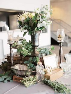 an arrangement of flowers and greenery on a table with wine glasses, silverware and candlesticks