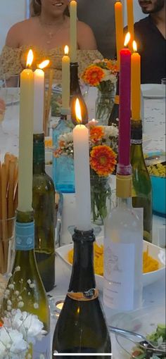 several candles are lit on the table with bottles and plates in front of them, as well as flowers