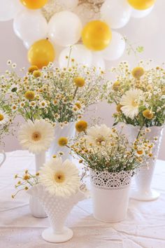 three white vases filled with yellow and white flowers on a table next to balloons