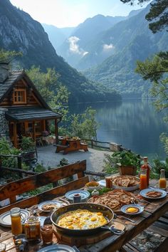 an outdoor table with food and drinks on it in front of a mountain lake surrounded by mountains