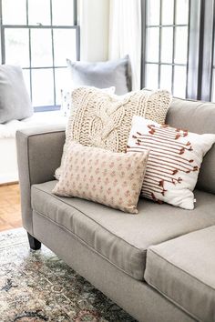 a couch with pillows on top of it in front of two windows and a rug