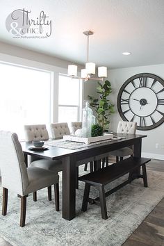 a dining room table with chairs and a large clock on the wall behind it in front of a window