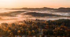 an aerial view of a town surrounded by trees in the fall with fog and low lying clouds