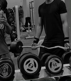 two men standing next to each other holding barbells and looking at their cell phones