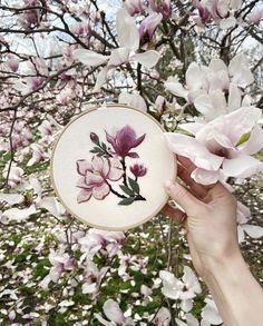 a person holding up a small embroidered plate with pink flowers on it in front of a flowering tree