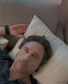 a man laying on top of a gray couch next to pillows and a remote control