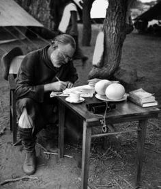 an old man sitting at a table writing in front of a tent with tents behind him