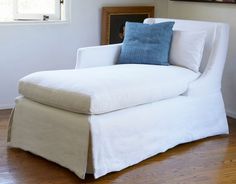 a white chaise lounge chair with blue pillows on it's back in front of a window