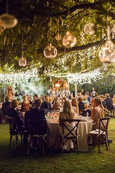 a group of people sitting around a table under a tree with lights hanging from it