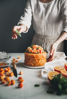 a woman is decorating a cake with oranges and other fruit on the table