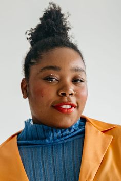 a woman wearing an orange jacket and blue shirt with her hair in a bun, looking at the camera