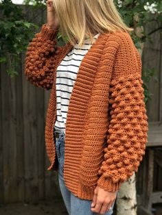 a woman standing in front of a tree wearing an orange cardigan sweater and jeans