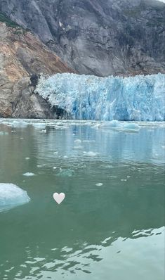 a heart shaped object floating in the water next to a large iceberg with mountains in the background