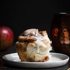 a cinnamon roll with icing on a plate next to an apple and two mugs