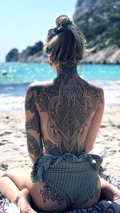 a woman sitting on top of a beach next to the ocean with tattoos on her back