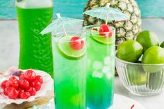 two glasses filled with green and red liquid next to pineapples on a plate