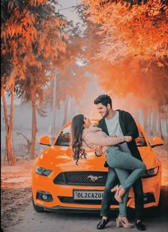 a man and woman sitting on the hood of an orange car with trees in the background