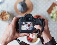 a person holding up a camera with food on the table in the back ground behind them