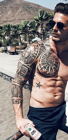 a man with tattoos on his arm and chest holding a skateboard near the beach