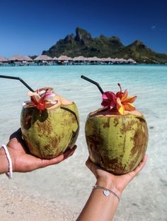 two coconuts with straws and flowers on them are held in front of the ocean